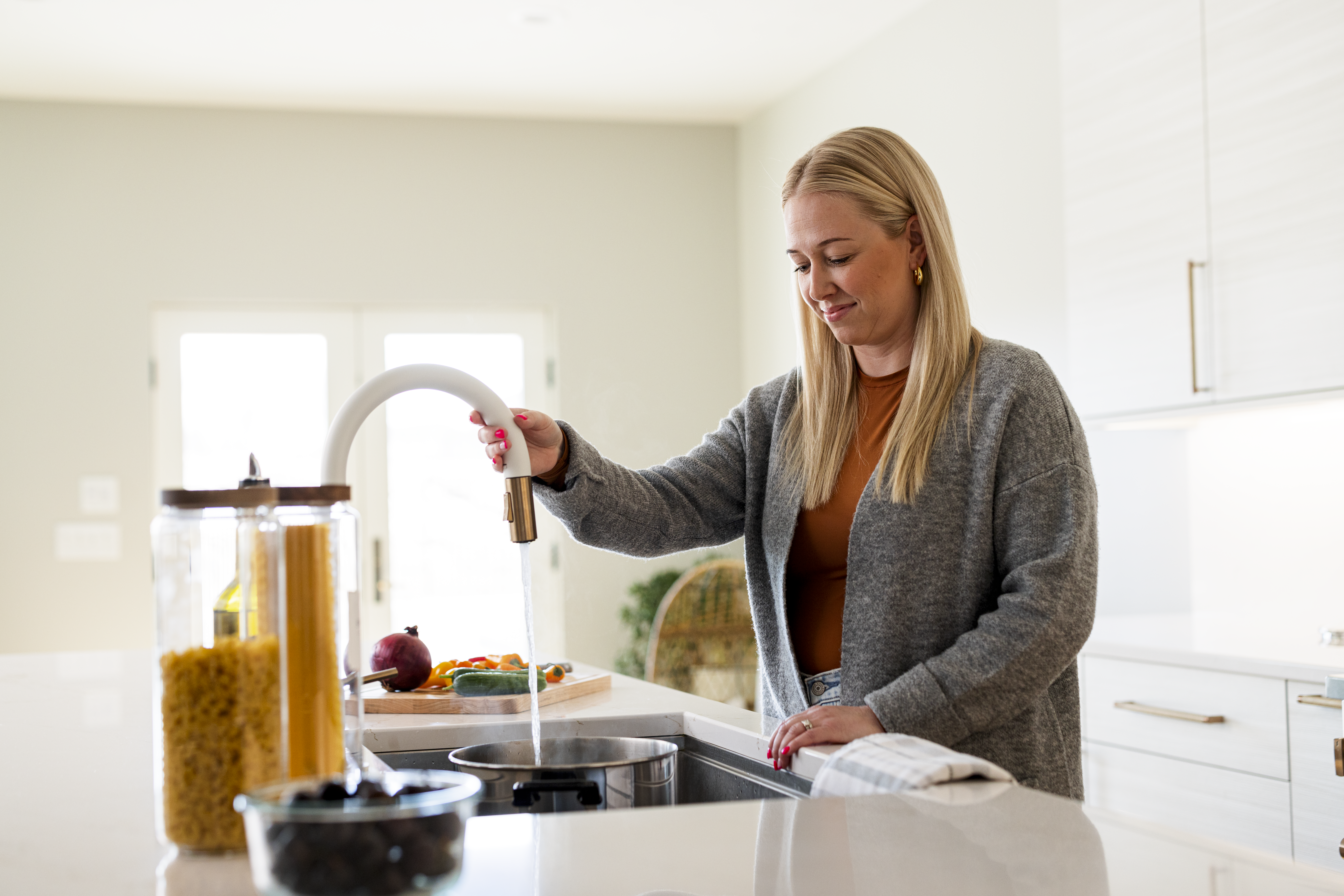 Woman filling a pot with water from a kitchen sink faucet, with fresh vegetables and pasta ingredients on the counter, preparing a meal in a bright modern kitchen.